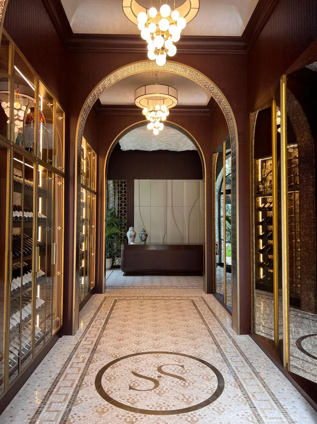 Elegant arched hallway with a beige marble mosaic floor and circular monogram medallion between gold-framed wine cabinets
